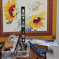 Wide view showing paint trim guide, caulk holder, black spirit level, blue caulking gun, assorted tools in containers with sunflower painting background