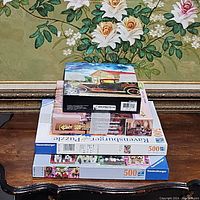 Four stacked puzzle boxes on a wooden table in front of floral wall art, showing puzzle themes and box condition.