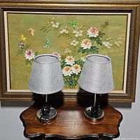 Pair of metal table lamps with grey textured shades placed on a wooden table in front of framed floral artwork.