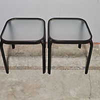 Two square patio side tables with black metal frames and frosted glass tops shown side by side on a concrete floor.