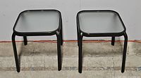 Two black metal frame square side tables with smoked glass tops shown side by side on a concrete floor against a white wall.