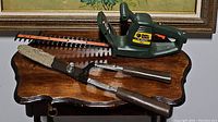 Black & Decker green electric hedge trimmer and gardening shears on a wooden table under a framed floral painting.