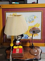 Photo showing vintage Mickey Mouse lamp with cream pleated shade, metal gooseneck desk lamp, and red untested push button telephone on wooden surface in front of floral artwork.