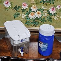 Both Brita UltraMax water container and blue Coleman cooler jug together on a wooden table with floral painting background