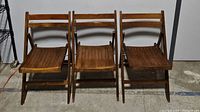 Three solid wooden folding chairs with slatted seats and backs, arranged side by side on a concrete floor.