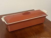 Side angled view of a terracotta rectangular Dutch oven with a matching lid and white handles with ridges.