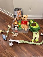 View of multiple stuffed animals including Winnie the Pooh, Tigger, penguin, and green lizard plush in a clear plastic bin. Also visible is the avocado costume with green hood and striped leggings, and the white plastic arm of the crib mobile with hanging soft toys on the floor.