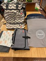Overview of the chevron Skip Hop diaper bag, stroller pocket, Mante stroller shield, Peekaboo baby packs, and foldable travel changing station on table.