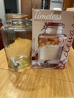 Clear glass drink dispenser with metal spigot sitting next to its original box showing product image.