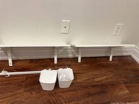 Two white wall shelves mounted on the wall with brackets, white hanging bar and two white hanging bins placed on floor