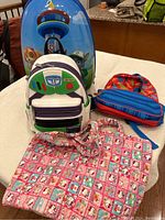 Set of five kids bags shown together on table: Paw Patrol rolling suitcase, Toy Story Buzz Lightyear mini backpack, blue and red backpack, Hello Kitty tote bag, and blue and red lunch bag.