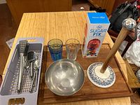 Photo showing assortment of kitchen utensils in a plastic basket, two drinking glasses, metal mixing bowl, box of candy, and decorative paper towel holder on wooden table.