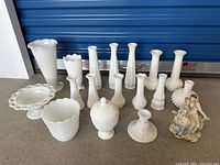 Wide view of 18 milk glass items including vases, plates, candlesticks, a covered dish, and a porcelain figurine, showing variety of shapes and textures.