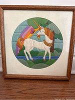 Full view of the framed needlepoint artwork depicting a unicorn with a rainbow and greenery background.