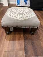 Front view of nursery footstool showing embroidered fabric top, nailhead trim, and dark wooden legs on wooden floor.