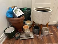 Collection of various indoor plant pots including large white ceramic pot on legs, green ceramic pot, terra cotta pots, decorative pots, and a glass terrarium box.