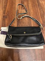 Front view of black leather purse with flap closure, gold Tory Burch logo and zipper detail on front pocket, resting on a cloth dust bag.
