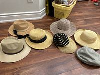 Seven different summer hats displayed on wood floor, varying in shape, size and color with ribbon accents and woven details.