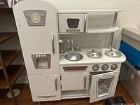 Front view of white play kitchen showing all main features including fridge, microwave, sink, stove and oven with oven door open.