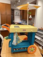 Full view of the Little Tikes lemonade and ice cream stand with umbrella, wheels, and a lemon print cloth hanging on the side.