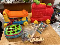 Wide shot showing all toys including VTech train, ride-on toy, shape sorter, wooden pull-along, and bags of assorted small toys and blocks on a table
