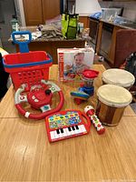 Lot overview showing all toys on a wooden table including shopping cart, steering wheel, drums, spinning top, toy piano, rattle, and boxed Hape String-along Shapes set.