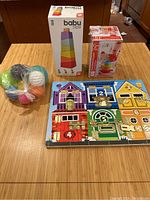 Wooden toys lot shown on table with all items visible: mesh bag with colorful balls, boxed wooden stacking toy, boxed wooden ring stacking toy, and Melissa & Doug latch board.
