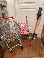 Photo showing metal pretend shopping cart, polka dot doll stroller, and toy stick vacuum together against a door background
