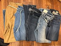 Seven pairs of women's jeans and pants laid flat on a wooden floor, showing color and styles including tan pants with drawstring and various denim washes.