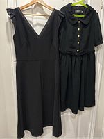 Full view of two black dresses hanging on wooden hangers, one sleeveless and one short sleeved with collar and buttons