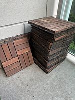 Stacked wooden balcony tiles showing interlocking edges and wood slat patterns, dark brown color.