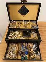 Front view of open black jewelry box showing divided compartments with mixed rings, earrings, necklaces, and bracelets visible