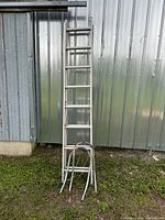 Aluminum extension ladder with extension legs shown against metal siding, standing vertically.