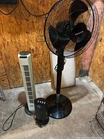 Three fans placed on a concrete floor against a wooden paneled wall; includes Honeywell pedestal fan, small black oscillating fan, and white tower fan.