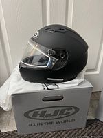 Matte black full face motorcycle helmet on top of gray box with HJC logo, showing clear visor and vents.