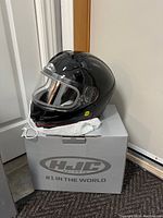 Black full-face motorcycle helmet with clear visor placed on top of an HJC branded helmet box.