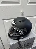 Black matte full-face motorcycle helmet with clear visor and HJC logo, placed on its original grey box in front of a white door.