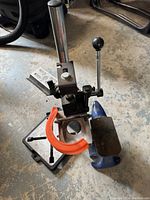 The photo shows a small metal anvil mounted on a base, a benchtop drill press with an adjustable platform and vertical column, and a large orange C-clamp attached to the drill press base on a concrete floor.