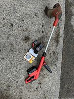 Photo of the red and black Black and Decker weed eater lying on the ground with two batteries and a charger placed beside it. Shows condition and included accessories.