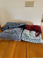Four folded blankets on wooden surface against white wall, including a blue queen size blanket in a bag, red plaid blanket, gray knit blanket, and blue and white patterned blanket.