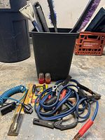 Photo shows a black container holding several car brushes and ice scrapers, multiple booster cables and clamps, coiled blue air hose, trailer light adapters, and other small automotive tools and accessories placed on the ground.