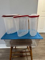 Three translucent plastic cereal containers with red lids arranged on a small blue table, along with a large clear food grade storage container underneath the table.