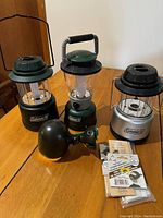Four Coleman battery-powered lanterns on a wooden table showing different models and colors