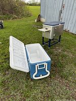 Two coolers outside on grass near a building: a large blue and white rolling plastic cooler with lid open, and a blue-silver metal cooler on stand behind it.