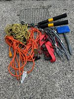 Photo showing tangled orange extension cords, hand pruners with black and yellow handles, red rubber bungee cords, a blue-handled hammer, metal gold-colored chain, and metal stakes.