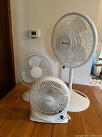 Photo shows three fans on a wooden table against light color wall, includes large Lasko pedestal fan, smaller Holmes oscillating fan, and smaller Holmes desk fan.