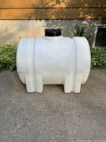Front view of the white plastic water fluid container tank with black screw-on lid, placed outside on concrete with plants and wall background.