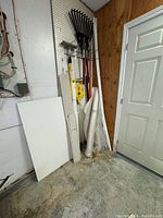 Photo of a garage corner showing two white rolls of vapor barrier plastic leaning against the wall alongside a black metal leaf rake and a yellow-black telescoping pole trimmer. Several white pieces of trim and poles are also visible.