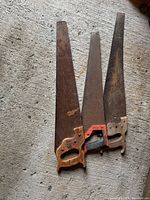 Three vintage hand saws laid on concrete floor, each showing rust and wear; one wooden handle, one red plastic handle, and one dark wooden handle visible