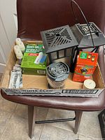 Older Therma Cell mosquito repellent products arranged in a white cardboard box on chair, visible items include black metal lanterns, boxed refill cartridges, OFF! torch fuel, assorted small parts and a tealight candle.